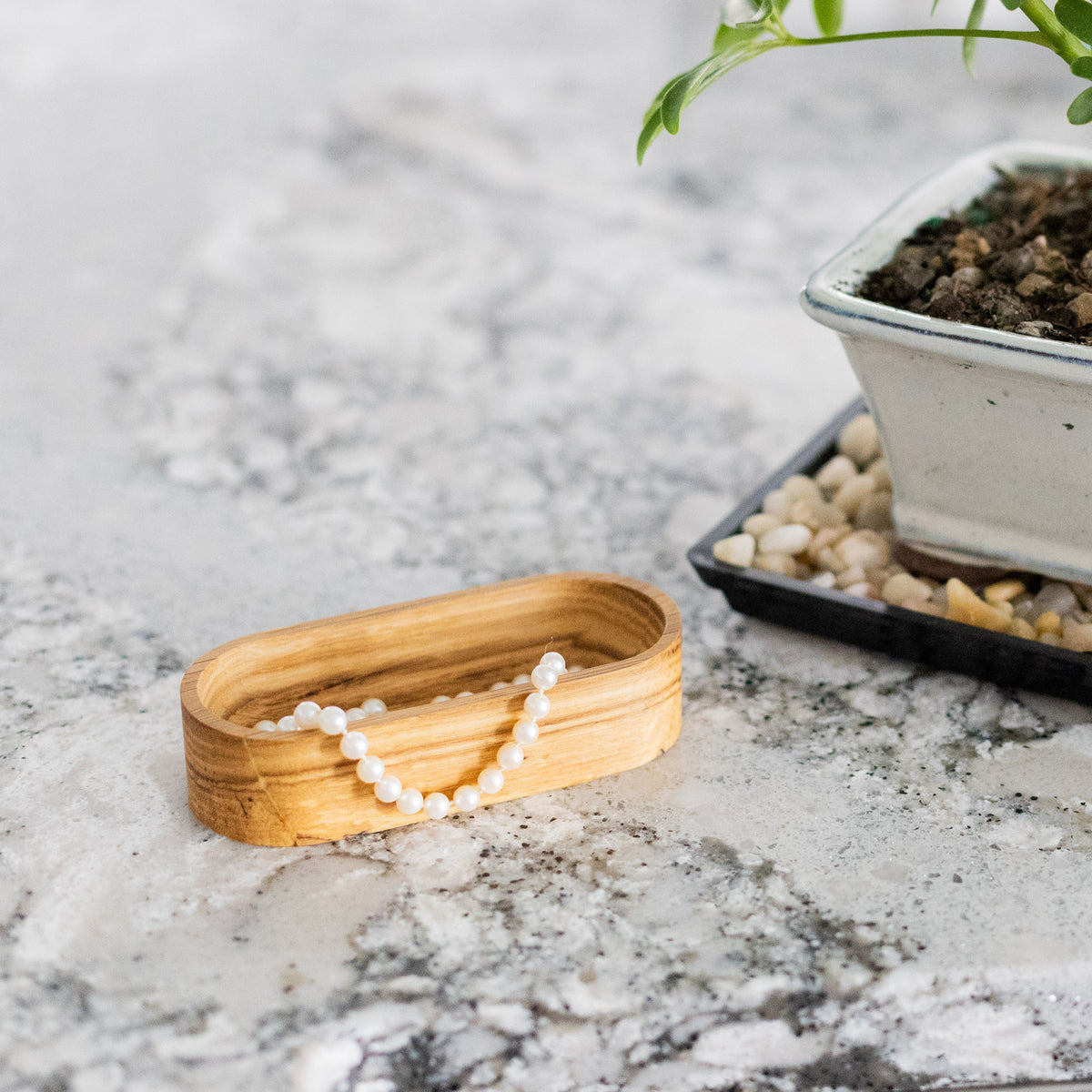 Solid oak 4 x 2 inch oval dish holding a necklace on a granite counter near a potted plant