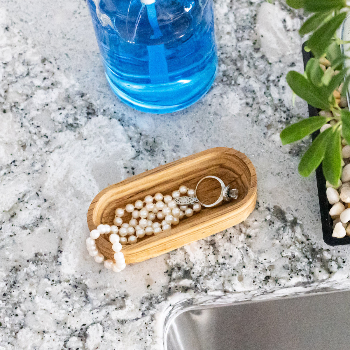 Solid oak oval ring dish holding a pearl necklace on a granite vanity beside a blue bottle